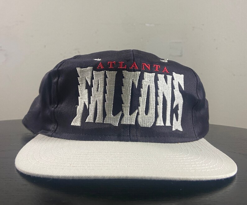 May include: A black and white baseball cap with the text "Atlanta Falcons" embroidered on the front in white with red accents.