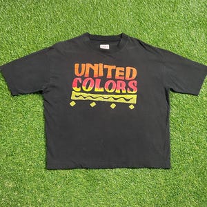 May include: Black t-shirt with the words "UNITED COLORS" in orange and red block letters. Below the text is a yellow wavy line with small yellow diamonds. The shirt is laid out on a green grassy surface.