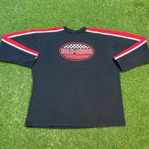 May include: Black long-sleeve t-shirt with red and white stripes on the sleeves. The shirt features a white oval with a black and white checkered pattern and the text "Harley-Davidson Motorcycles" in red.