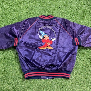 May include: A purple satin bomber jacket with a red and blue striped trim. The jacket features an embroidered image of Mickey Mouse dressed as a magician.