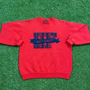 Könnte beinhalten: Roter Pullover mit einem rot-marineblauen Karomuster, auf dem in einem Banner "Holy Family" steht.