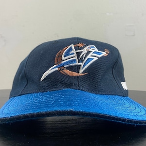 Puede incluir: Una gorra de béisbol negra y azul con un logotipo bordado en blanco y naranja de un jugador de baloncesto con una estrella y una luna creciente. El logotipo está en la parte delantera de la gorra.
