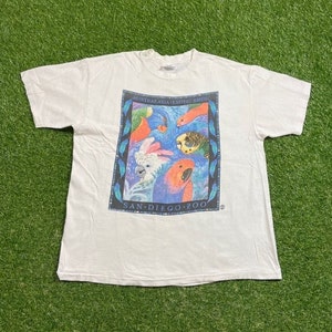 May include: White t-shirt featuring a colorful graphic of exotic birds, including parrots and cockatoos, within a blue-framed design. The text "AUSTRALASIA EXOTIC BIRDS" and "SAN DIEGO ZOO" are printed on the shirt.