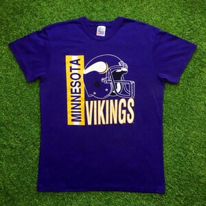 May include: Purple t-shirt with a yellow and white Minnesota Vikings logo featuring a football helmet and the words "Minnesota Vikings".