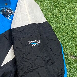 Peut inclure: Veste Reebok vintage bleue, noire et blanche. La veste pr&eacute;sente le logo des Panthers de la Caroline sur la poitrine et le logo Reebok sur la manche. La veste est en nylon.