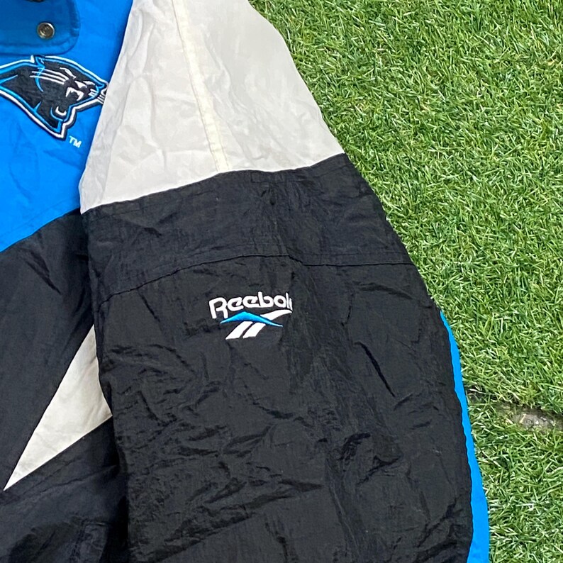 Peut inclure: Veste Reebok vintage bleue, noire et blanche. La veste pr&eacute;sente le logo des Panthers de la Caroline sur la poitrine et le logo Reebok sur la manche. La veste est en nylon.