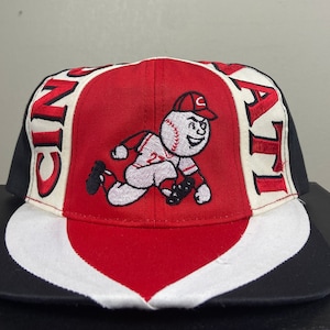 May include: A red, white and black baseball cap with the word "CINC" on the left side and "ALT" on the right side. A cartoon baseball player with a baseball for a head is embroidered in the centre of the cap.