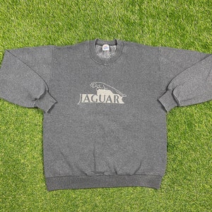 Puede incluir: Una sudadera gris oscuro con el logotipo de Jaguar en gris claro. El logotipo presenta un jaguar saltando sobre el nombre de la marca. La sudadera tiene cuello redondo y mangas largas con puños acanalados.