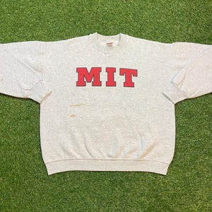 Puede incluir: Una sudadera gris claro con las letras rojas "MIT" en el pecho. La sudadera tiene mangas largas y cuello redondo. La tela parece ser una mezcla de algodón.