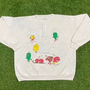Könnte beinhalten: Weißer Langarm-Pullover mit Druckknopfverschluss am Hals. Der Pullover zeigt ein farbenfrohes Cartoon-Design mit einer Biene, einer Zitrone, Kirschen und grünen Figuren.