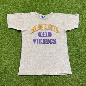 May include: A gray Champion t-shirt featuring the text "MINNESOTA" in gold arched lettering above an oval with "XXL" in purple, and "VIKINGS" in purple below. The shirt is on a green surface.