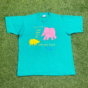 Op de afbeelding: Een turquoise blauw t-shirt met een grafische print van een roze olifant, een gele neushoorn en een blauwe walvis. De tekst op het shirt luidt "animals in danger now you see us... soon you won't".