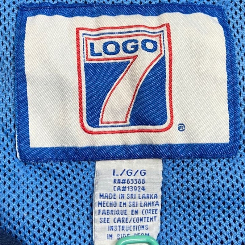 Peut inclure: Une &eacute;tiquette de v&ecirc;tement bleue et blanche avec le texte "LOGO 7" en rouge et blanc. L'&eacute;tiquette comprend &eacute;galement le texte "L/G/G", "RN#63388", "CA#13924", "MADE IN SRI LANKA", "MECHO EM SRI LANKA", "FABRIQUE EN COREE", "SEE CARE/CONTENT INSTRUCTIONS IN SIDE SEAM".