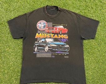 Op de afbeelding: Zwart T-shirt met een afbeelding van een Ford Mustang. De afbeelding bevat de tekst "Ford Mustang" en de jaren "1964 1/2 - 1993" en "1994 GT 5.0".