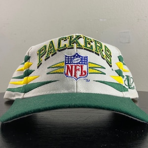 Puede incluir: Gorra de béisbol blanca y verde con las palabras "Packers" y "NFL" bordadas en la parte delantera. La gorra tiene una visera verde y rayas amarillas y verdes en los lados.