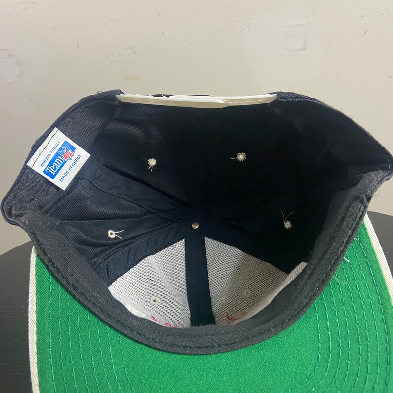 May include: Black and green baseball cap with a white brim and a white mesh lining. The cap has a black label on the inside that says "Team Up" and "One Size Fits All" with "Made in China" printed below.