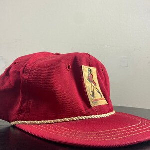 Op de afbeelding: Een rode baseballpet met een witte touwrand en een patch met een persoon in een rood uniform met de tekst "Johnnie Walker Red Label" op de patch.
