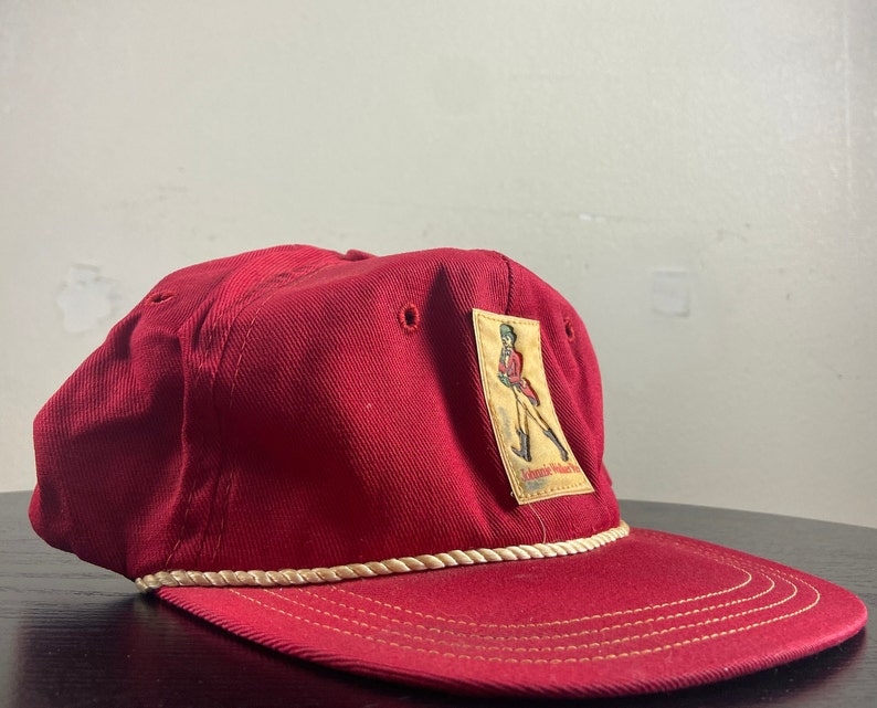 Op de afbeelding: Een rode baseballpet met een witte touwrand en een patch met een persoon in een rood uniform met de tekst "Johnnie Walker Red Label" op de patch.
