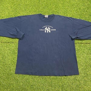 May include: A navy blue long-sleeve t-shirt featuring a white graphic on the chest. The graphic displays the New York Yankees logo in the center, flanked by the text Yankee Stadium and the years 1923 and 2008 printed in a white circular arc.