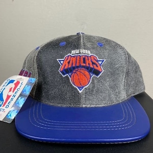 Puede incluir: Una gorra de béisbol gris y azul con el logotipo de los New York Knicks. El logotipo es una pelota de baloncesto azul y naranja con las palabras "New York Knicks" en un arco azul y blanco sobre la pelota. La gorra tiene una visera azul y una corona gris. La gorra tiene una etiqueta que dice "Producto oficial con licencia de la NBA" con el número 206464573.