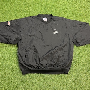 May include: A black long-sleeve pullover windbreaker jacket featuring a small white Philadelphia Eagles logo on the upper left chest, a crew neckline, elastic cuffs, and a small rectangular logo patch on the right sleeve near the shoulder.