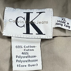 Pu&ograve; includere: Primo piano di un'etichetta Calvin Klein Jeans. L'etichetta bianca presenta testo nero e un grande logo "CK". L'etichetta indica che il capo &egrave; XL, prodotto a Hong Kong, ed elenca la composizione del tessuto come 60% cotone e 40% poliuretano.