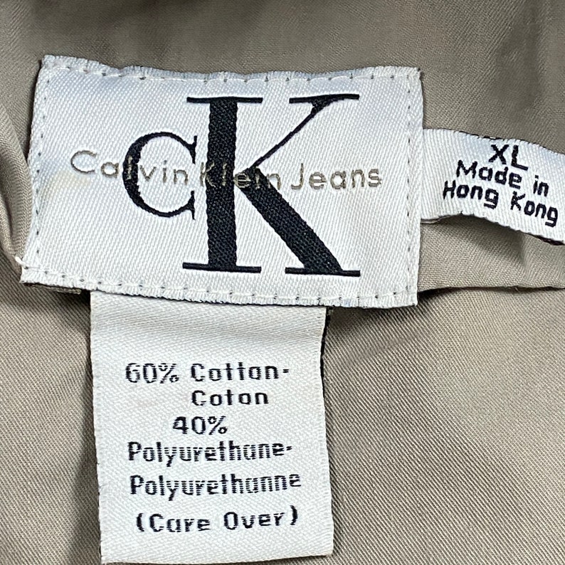 Pu&ograve; includere: Primo piano di un'etichetta Calvin Klein Jeans. L'etichetta bianca presenta testo nero e un grande logo "CK". L'etichetta indica che il capo &egrave; XL, prodotto a Hong Kong, ed elenca la composizione del tessuto come 60% cotone e 40% poliuretano.