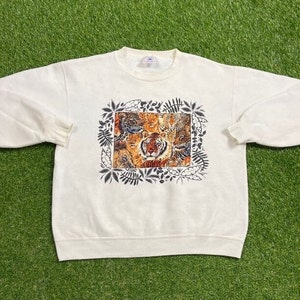 May include: A white crewneck sweatshirt with a graphic print of a group of tigers and leopards in a jungle setting. The print is surrounded by a black outline of leaves and vines.