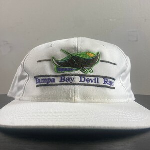 May include: White baseball cap with a black, green, and purple embroidered Tampa Bay Devil Rays logo featuring a manta ray. The text "Tampa Bay Devil Rays" is embroidered in purple and black.