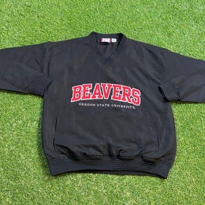 May include: Black pullover jacket with a V-neck and long sleeves. The jacket features the word "BEAVERS" in red block letters with a white outline, and the words "OREGON STATE UNIVERSITY" in smaller red letters.