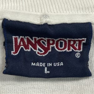 Może przedstawiać: Zbliżenie granatowej metki z nazwą marki "JANSPORT" w czerwono-białych literach. Poniżej nadruk "MADE IN USA". Etykieta jest przyszyta do białej tkaniny, a rozmiar "L" jest nadrukowany.