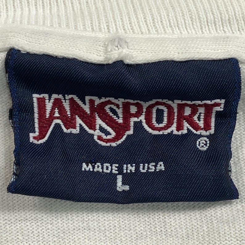 Może przedstawiać: Zbliżenie granatowej metki z nazwą marki "JANSPORT" w czerwono-białych literach. Poniżej nadruk "MADE IN USA". Etykieta jest przyszyta do białej tkaniny, a rozmiar "L" jest nadrukowany.