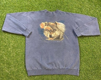 May include: A blue sweatshirt with a wolf graphic. The wolf is shown with detailed fur and is partly hidden by autumn leaves. The sweatshirt has long sleeves and a crew neck.