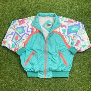 May include: A turquoise blue windbreaker jacket with a white, pink, and blue geometric pattern on the sleeves. The jacket has a zipper closure and a pink zipper pull. The text "River Edge" is printed on the front of the jacket.
