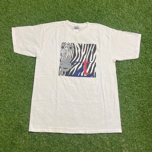 May include: White t-shirt with a zebra graphic. The zebra has black and white stripes, a grey muzzle, and a blue and red letter "t" design. The shirt is made of a soft, comfortable material.