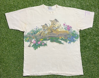 May include: A white t-shirt with a graphic print of two cougars in a forest setting. The cougars are surrounded by flowers and a hummingbird. The text "Cougar Mountain" is printed on the right sleeve.