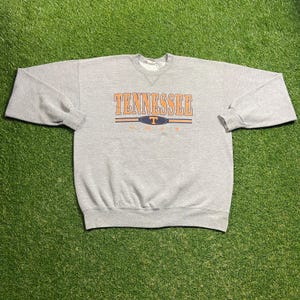May include: A gray sweatshirt with the word "TENNESSEE" in orange block letters across the chest. Below the word "TENNESSEE" is a blue oval with a white "T" inside. The word "VOLS" is below the oval. The sweatshirt is on a green surface.