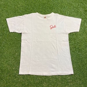 以下が含まれることがあります： 半袖の白いTシャツ。 「Stoli」の文字が赤い筆記体で胸の左上にプリントされています。 シャツは柔らかい素材で作られています。