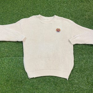 以下が含まれることがあります： クリーム色のクルーネック長袖ニットセーター。胸元に小さな刺繍エンブレムが付いています。セーターは厚手のテクスチャードニット生地で作られています。