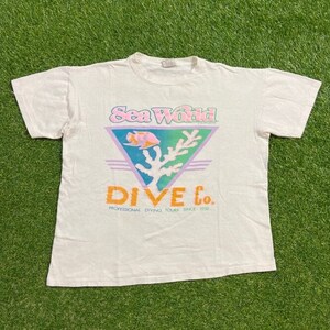 Puede incluir: Camiseta blanca con un logotipo colorido de Sea World Dive Co. El logotipo presenta un pez rosa y naranja, un arrecife de coral verde y blanco, y el texto "Sea World Dive Co. Professional Diving Tours Since 1988."