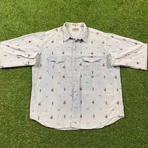 Puede incluir: Una camisa de manga larga azul claro con botones y un patrón repetido de vaqueros montando toros. La camisa tiene dos bolsillos en el pecho y una etiqueta blanca con el texto "L.L.Bean" en el lado izquierdo del cuello.