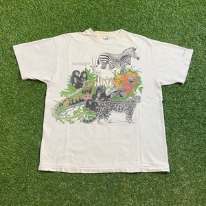 May include: Off-white t-shirt featuring a colorful graphic of various animals, including a zebra, lion, and leopard. The design incorporates a green iguana, monkeys, and other wildlife. The shirt has short sleeves and a crew neck.