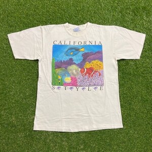 以下が含まれることがあります： 白いTシャツ。カラフルな水中海の生物のグラフィック、魚やサンゴなど。画像の上と下に「CALIFORNIA STYLE」の文字がプリントされています。柔らかく快適な素材で作られています。