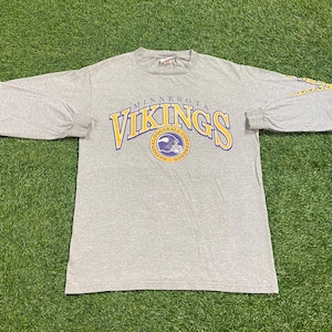 May include: A heather gray long-sleeve t-shirt featuring the Minnesota Vikings logo in yellow and purple. The logo includes the team name and a football helmet. The shirt is laid flat on a green surface.