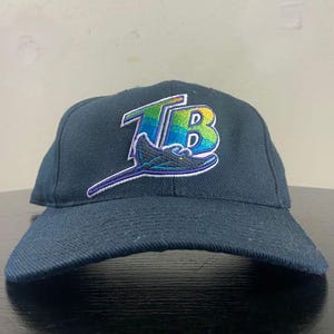 May include: A navy blue baseball cap featuring the Tampa Bay Rays logo. The logo is an embroidered design with the letters "TB" in green, blue, and yellow, with a stylized ray. The cap has a curved brim and a structured crown.