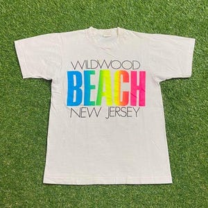 May include: White t-shirt with the words "WILDWOOD BEACH NEW JERSEY" in colorful block letters. The word "BEACH" is in a rainbow gradient. The shirt is on a green grassy surface.