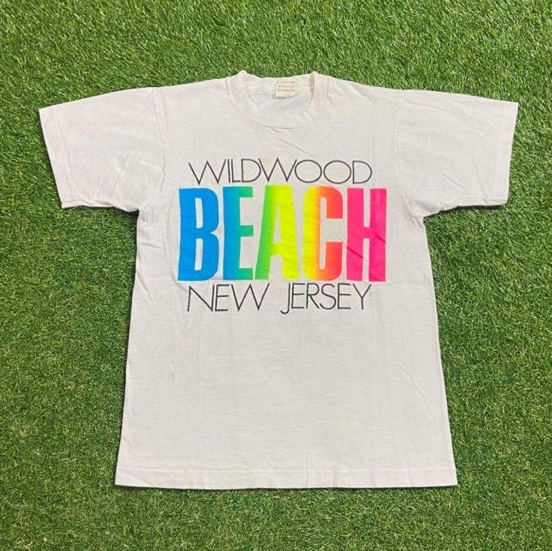 May include: White t-shirt with the words "WILDWOOD BEACH NEW JERSEY" in colorful block letters. The word "BEACH" is in a rainbow gradient. The shirt is on a green grassy surface.