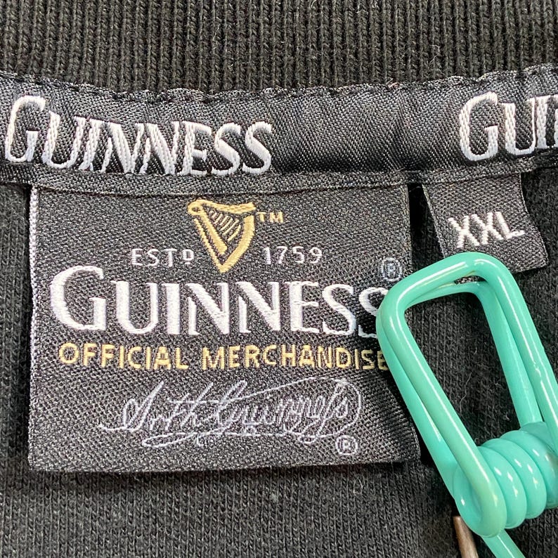 May include: Black t-shirt label featuring the Guinness logo, a gold harp, and the text "ESTD 1759". The label also reads "OFFICIAL MERCHANDISE" and "Arth Guinness". The size is XXL.