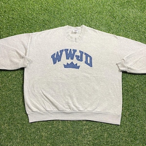 Könnte beinhalten: Hellgraues Sweatshirt mit dem blauen Text "WWJD" in Bogenform über einer blauen Krone. Das langärmlige Oberteil mit Rundhalsausschnitt ist aus einem weichen Material gefertigt. Das Kleidungsstück liegt auf einer grünen Oberfläche.