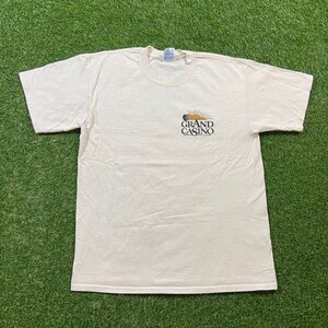 May include: A white t-shirt with a graphic of a sun setting behind a building with the text "Grand Casino" printed on it.